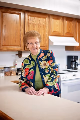 An elderly woman with glasses and a colorful floral jacket stands in a kitchen, leaning on a countertop. The kitchen has wooden cabinets, a white stove, a coffee maker, and containers on the counter.
