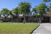Single-story building with a brown roof and beige walls, surrounded by green grass and several trees under a clear blue sky. A concrete walkway leads to the entrance of the building.