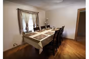 A dining room with a rectangular table covered by a beige tablecloth and eight dark wooden chairs. The table is set with placemats and a centerpiece. There is a window with curtains on the left wall and a lamp in the corner. The floor is wooden and the walls are light-colored.