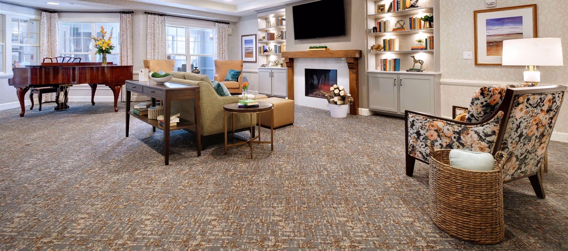 A cozy living room area with a patterned carpet, a grand piano with a vase of flowers on top, a beige sofa with green pillows, two tan armchairs, a round side table with books and a small flower vase, a fireplace with a wooden mantle and a bucket of firewood, built-in shelves with books and decorative items, a wall-mounted TV above the fireplace, a floral upholstered chair next to a woven basket with a cushion, and a table lamp on a side table.