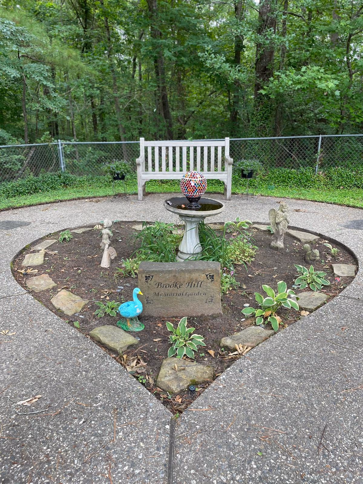 A heart-shaped garden area with a stone plaque that reads 'Brooke Hill Memorial Garden' surrounded by small plants and decorative statues including a blue duck and angels. In the center of the garden is a birdbath with a colorful mosaic ball on top. Behind the garden is a white wooden bench and a chain-link fence with dense green trees in the background.