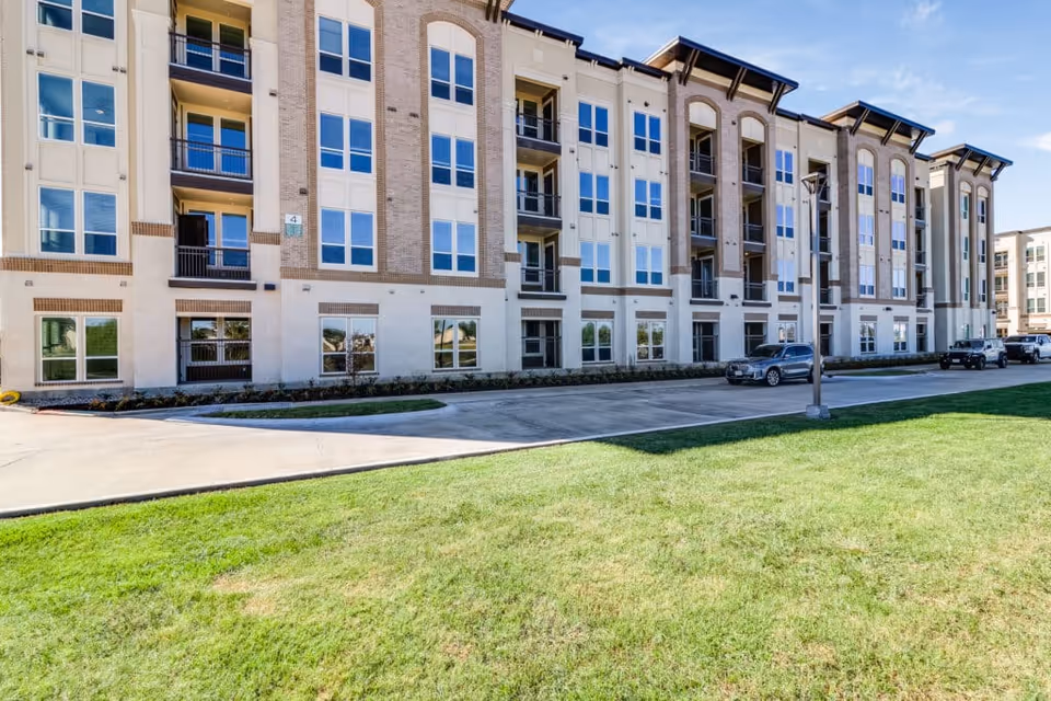 Front exterior of a multi-story senior living building with balconies, parked cars, and a grassy lawn.