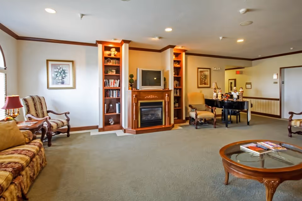 A cozy living room area in a senior living facility featuring a fireplace with a TV on top, built-in bookshelves on either side, comfortable armchairs, a sofa, a round glass coffee table with magazines, and a piano with decorative figurines. The room has beige walls with wood trim and carpeted flooring.