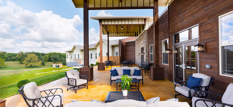 Outdoor covered patio area at Webwood Assisted Living with cushioned chairs and sofas arranged around a coffee table with a potted plant. The patio overlooks a green lawn and trees under a partly cloudy sky.