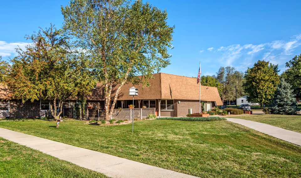 One-story brick senior living building with a fenced patio, flagpole, and a grassy lawn with trees under a blue sky.