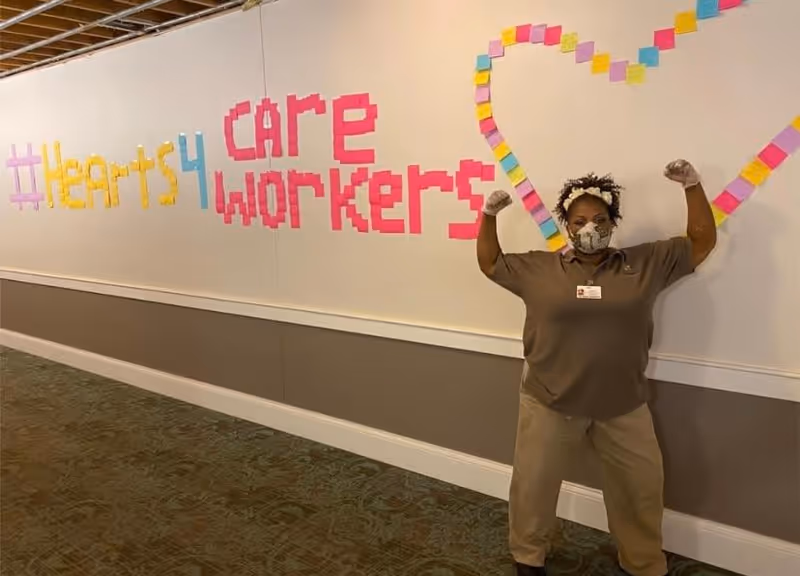 A person wearing a face mask and gloves stands in a hallway flexing their arms in a strong pose. Behind them on the wall, colorful sticky notes form the words '#hearts4 care workers' and a large heart shape.