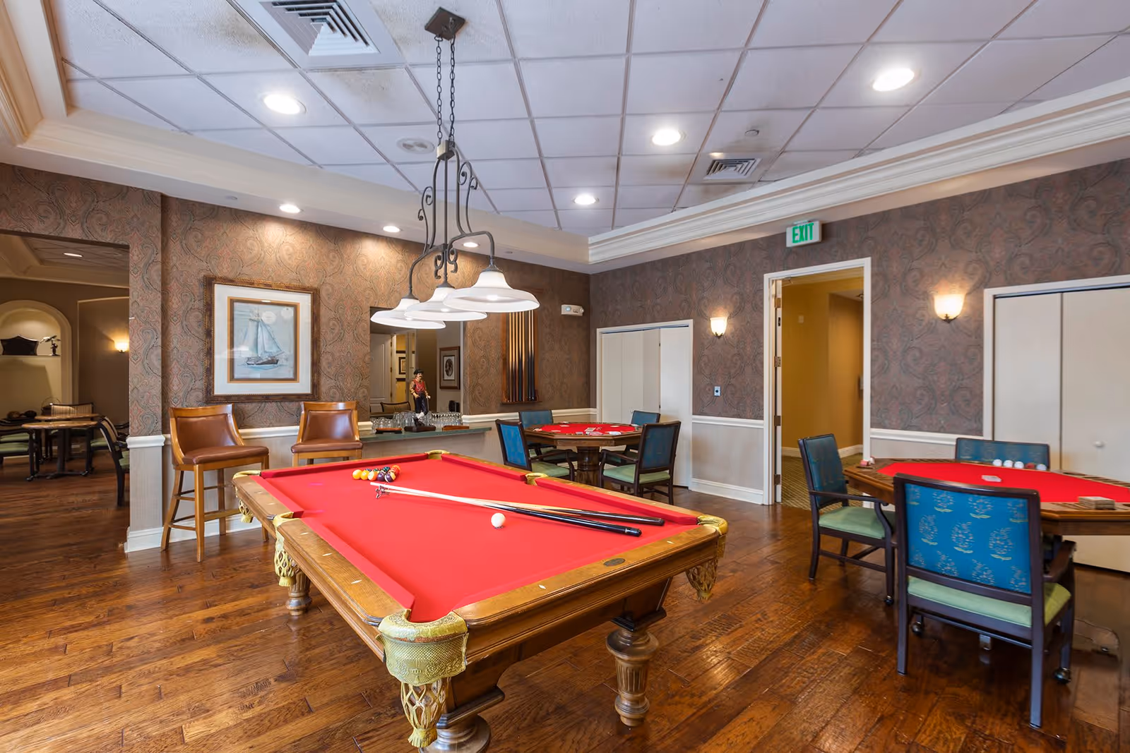 A game room with a red felt pool table in the center, pool cues and balls arranged on it. The room has wooden flooring, patterned wallpaper, and ceiling lights. There are two brown leather chairs against the wall, and two card tables with blue and green chairs around them. A framed picture of a sailboat hangs on the wall.