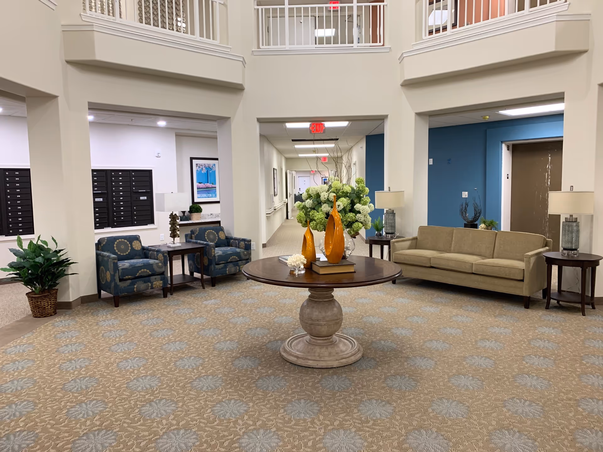 Lobby seating area with a round centerpiece table holding floral arrangements, surrounded by chairs and a sofa under a two-story ceiling.