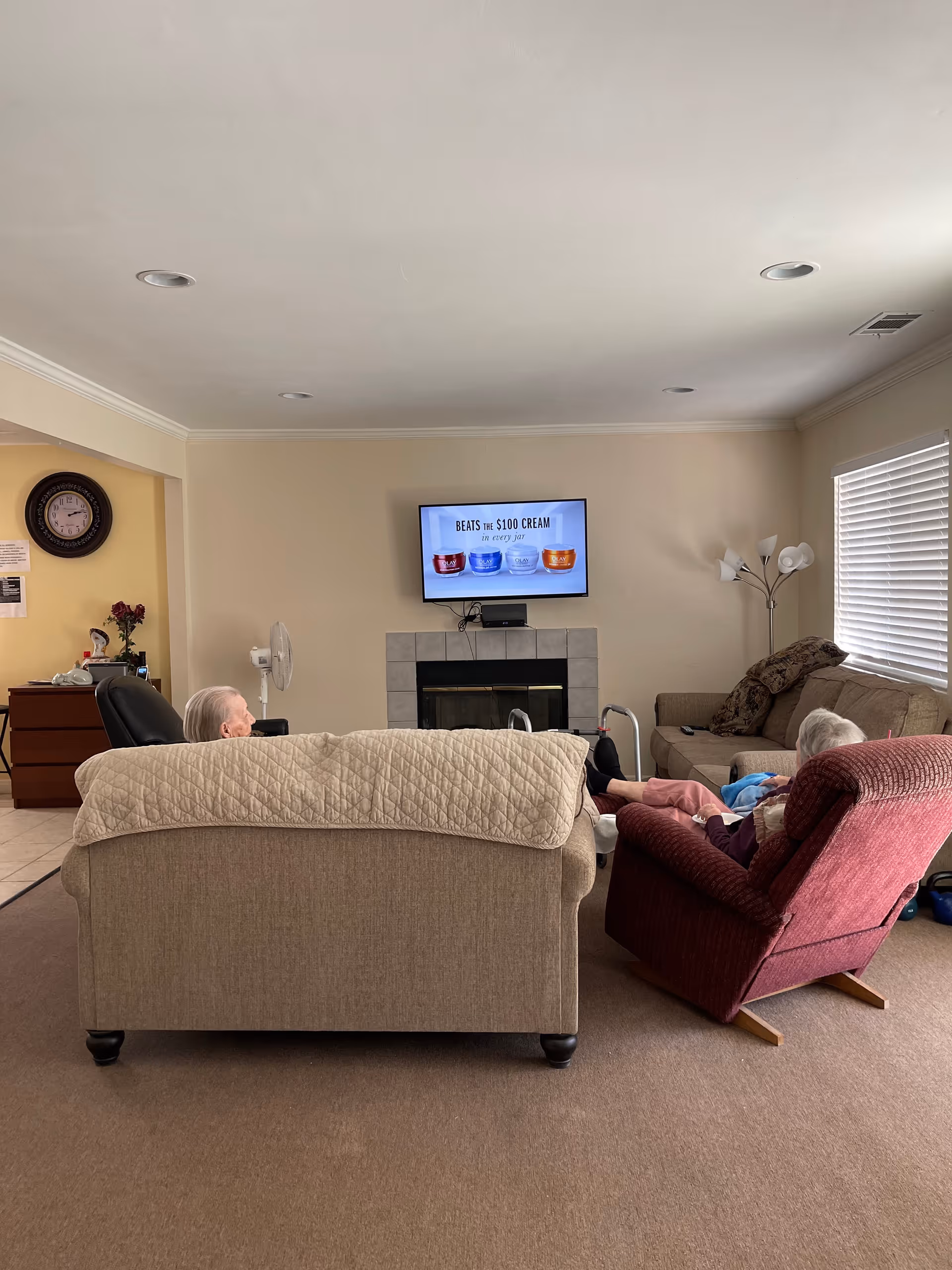 Living room with a couch and recliners facing a wall-mounted TV above a fireplace, two people seated, a floor lamp, and a window with blinds.