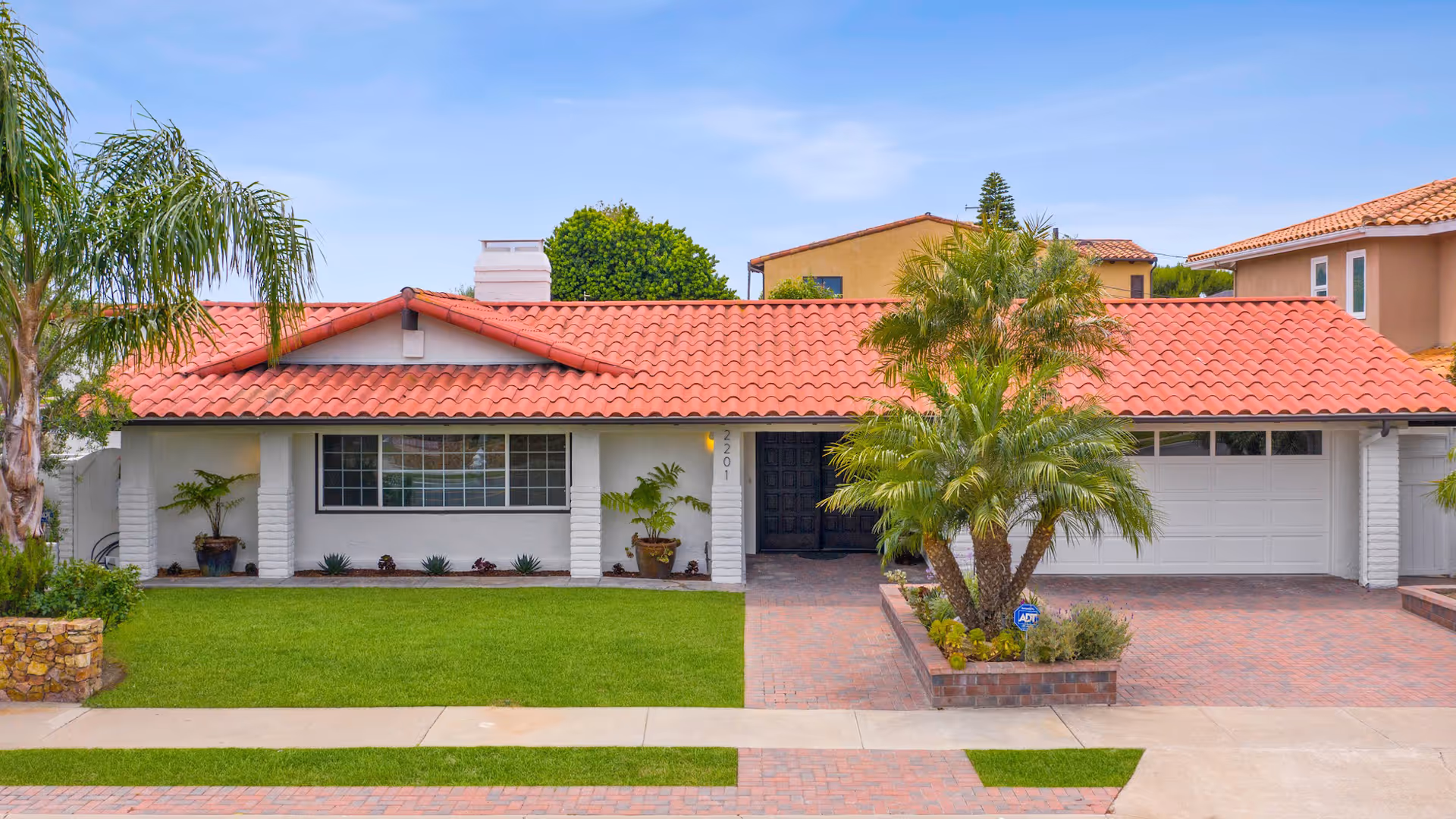 Single-story Spanish-style home with a red tile roof, attached garage, palm trees, and a manicured front lawn.