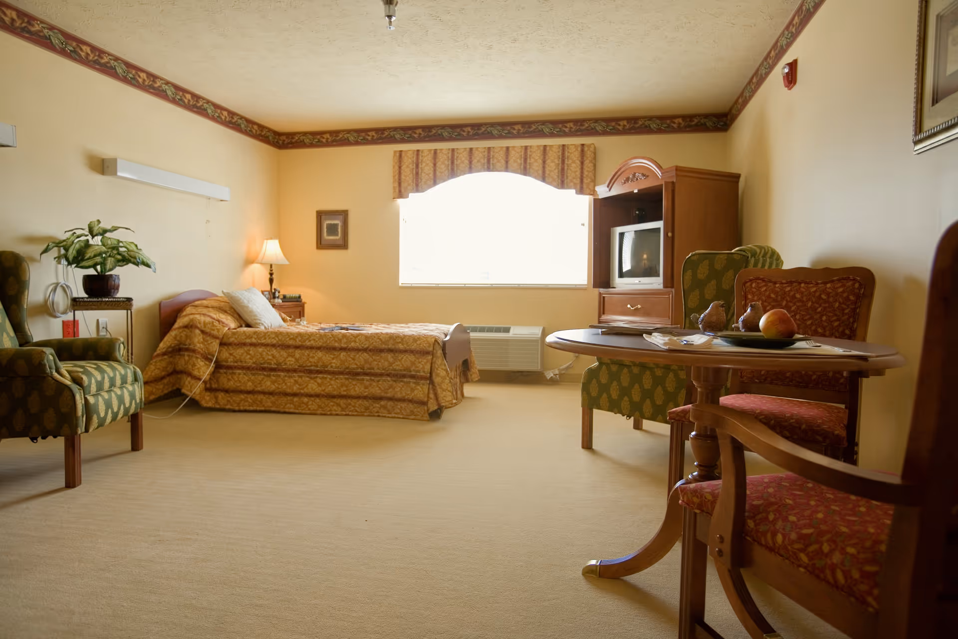 A cozy senior living room with a single bed covered in a patterned bedspread, a nightstand with a lamp, two upholstered armchairs, a wooden table with two chairs, and a wooden TV cabinet with an old-style television. The room has beige walls with a decorative border near the ceiling and a large window with a valance letting in natural light.