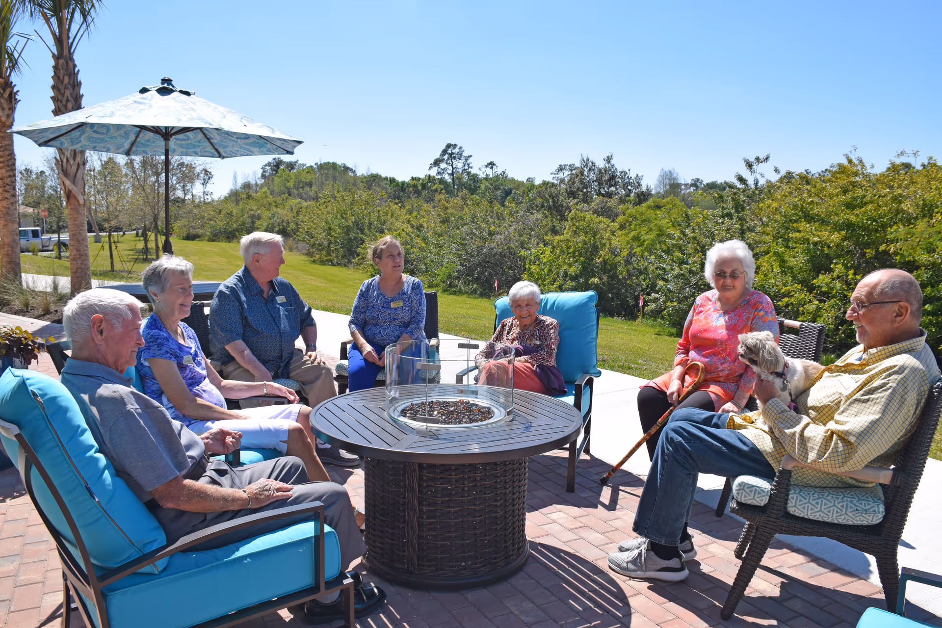 A group of elderly people sitting outdoors around a circular fire pit table on a patio. They are seated on cushioned chairs under a large umbrella, enjoying a sunny day with greenery and trees in the background. One man is holding a small dog on his lap.