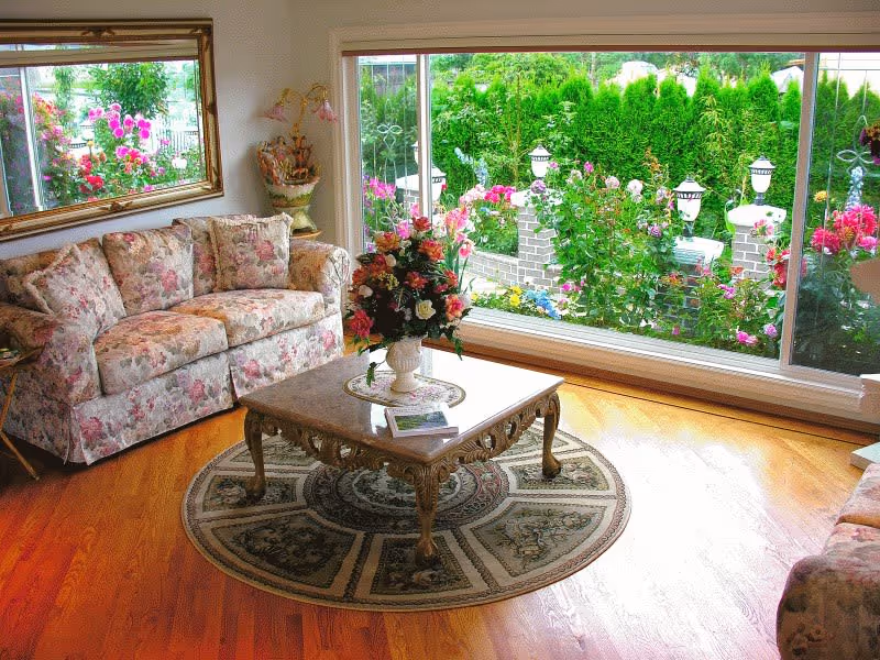 Sunlit living room with floral-patterned sofas, an ornate coffee table on a round rug, and a large window overlooking a blooming garden.