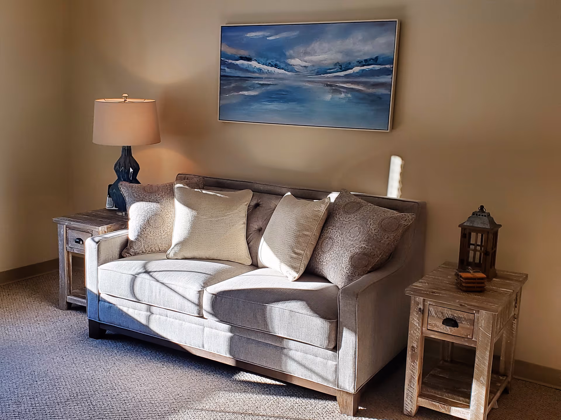 A cozy living room corner with a grey loveseat, patterned throw pillows, wooden side tables, a table lamp, and a seascape painting on the wall.