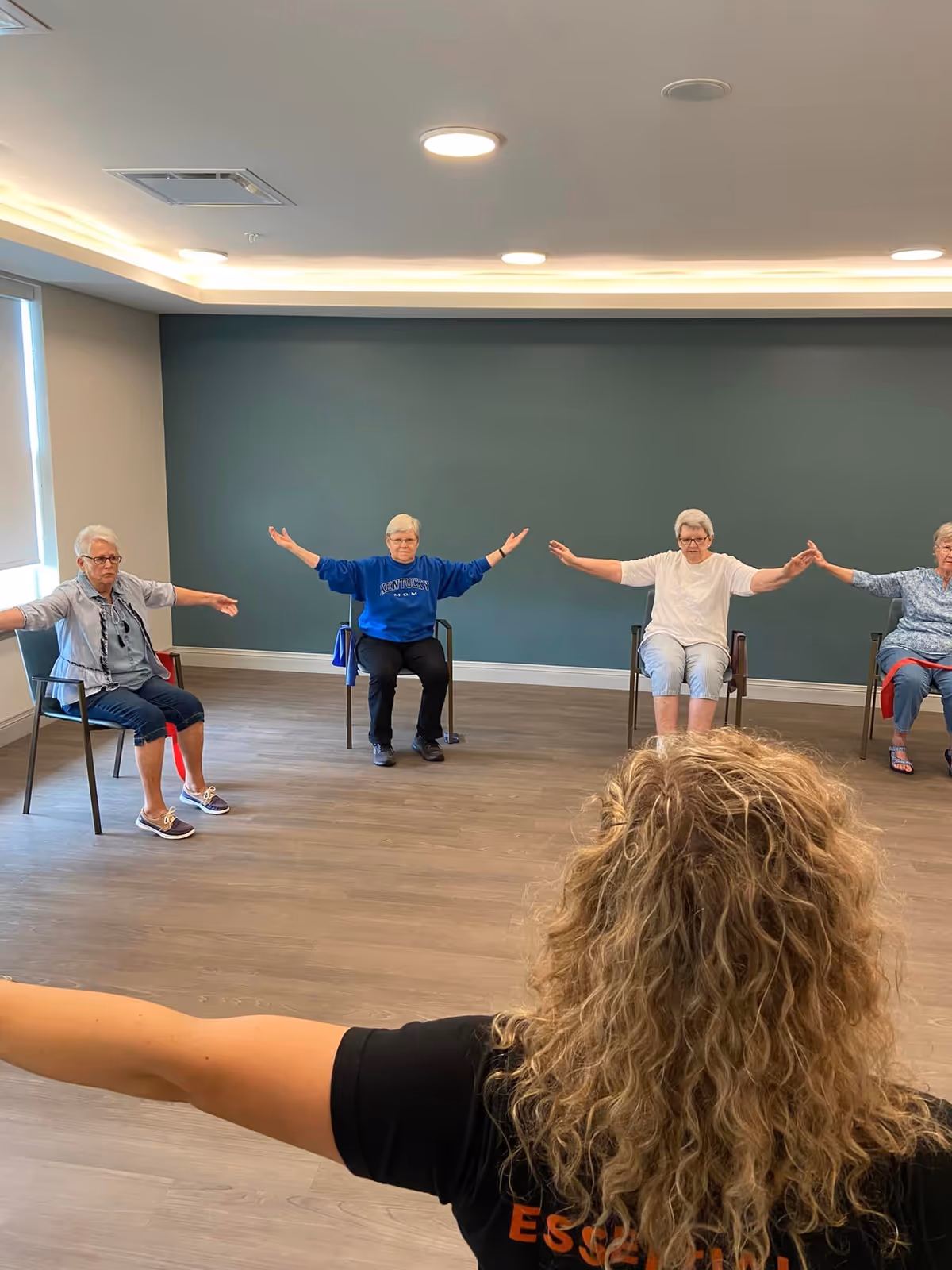 A group of elderly women seated in chairs in a spacious room with wooden flooring and a teal accent wall, participating in a seated exercise class led by an instructor with curly blonde hair wearing a black shirt.