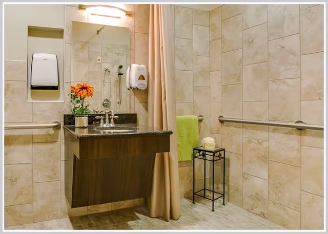 Wheelchair-accessible tiled bathroom with a wall-mounted sink and granite countertop, grab bars, shower curtain, and a towel on a small stand.