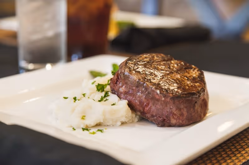 A close-up of a plated meal featuring a grilled steak and a serving of mashed potatoes garnished with chopped herbs, with a glass of iced beverage and water in the background.