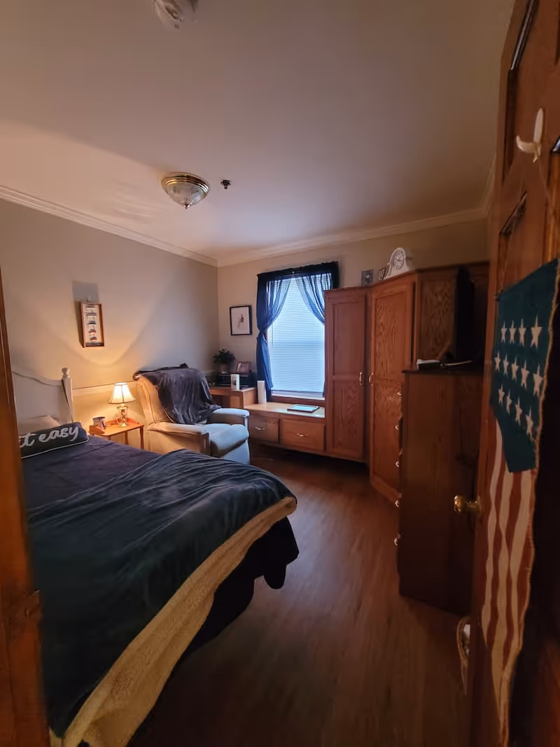 A cozy bedroom with a bed covered in dark blue and beige blankets, a cushioned armchair with a throw blanket, a wooden nightstand with a lamp, a window with blue curtains, and wooden furniture including a dresser and a cabinet.