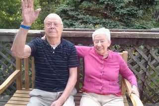 An elderly man and woman sitting on a wooden bench outdoors. The man is wearing a black striped polo shirt and beige pants, raising his right hand. The woman is wearing a pink cardigan and beige pants, smiling with her arm around the man's shoulder. Behind them is a wooden lattice fence and green trees.