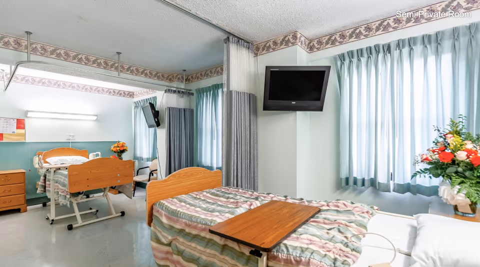 A semi-private hospital room with two beds separated by a privacy curtain. Each bed has a wooden headboard and footboard, with striped bedding. There is a wooden bedside table with a vase of flowers next to one bed, and a small wooden table on wheels over the other bed. Two wall-mounted televisions are visible, one near each bed. The room has large windows with light blue curtains and floral wallpaper border near the ceiling.