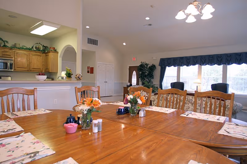 A spacious dining area with a large wooden table set with floral placemats, salt and pepper shakers, and small flower vases. In the background, there is a kitchen with wooden cabinets and a microwave, and a living room area with armchairs and large windows with blue valances letting in natural light.