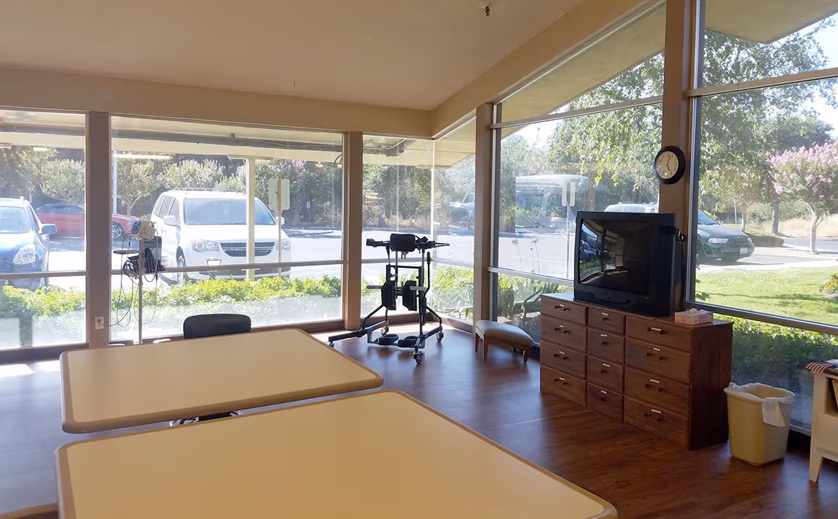 Interior view of a room with large windows showing a parking lot outside. Inside, there are two tables with beige tops, a wooden dresser with a TV on top, a clock on the wall, a walker, and a trash can. The room has wooden flooring and is well-lit with natural light.