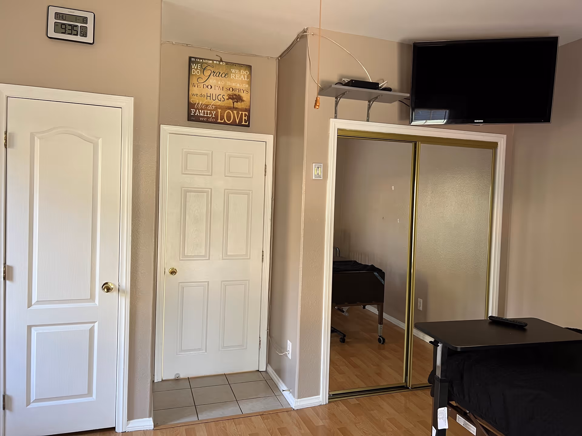 Interior view of a senior living bedroom with two white doors, a mirrored sliding closet, wall-mounted TV, and a hospital bed with an overbed table.