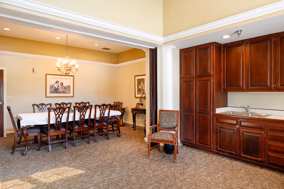 A formal dining room with a long table and multiple wooden chairs under a chandelier, adjacent to a kitchenette with dark wood cabinets and a sink.