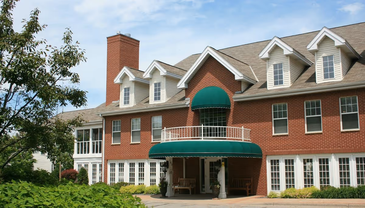 Exterior view of a large brick building with white-framed windows and a green awning over the entrance. The building has multiple dormer windows on the roof and a chimney. There are benches near the entrance and greenery including bushes and a tree in the foreground.