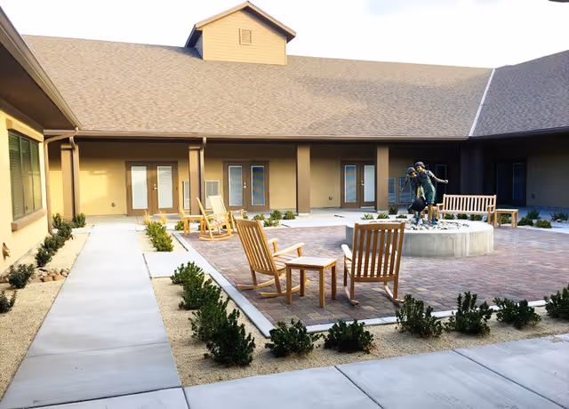 Outdoor courtyard area with a paved seating space featuring wooden chairs and a bench surrounding a circular fountain with a sculpture of two children playing. The courtyard is bordered by a building with multiple doors and windows, and landscaped with small bushes and gravel.