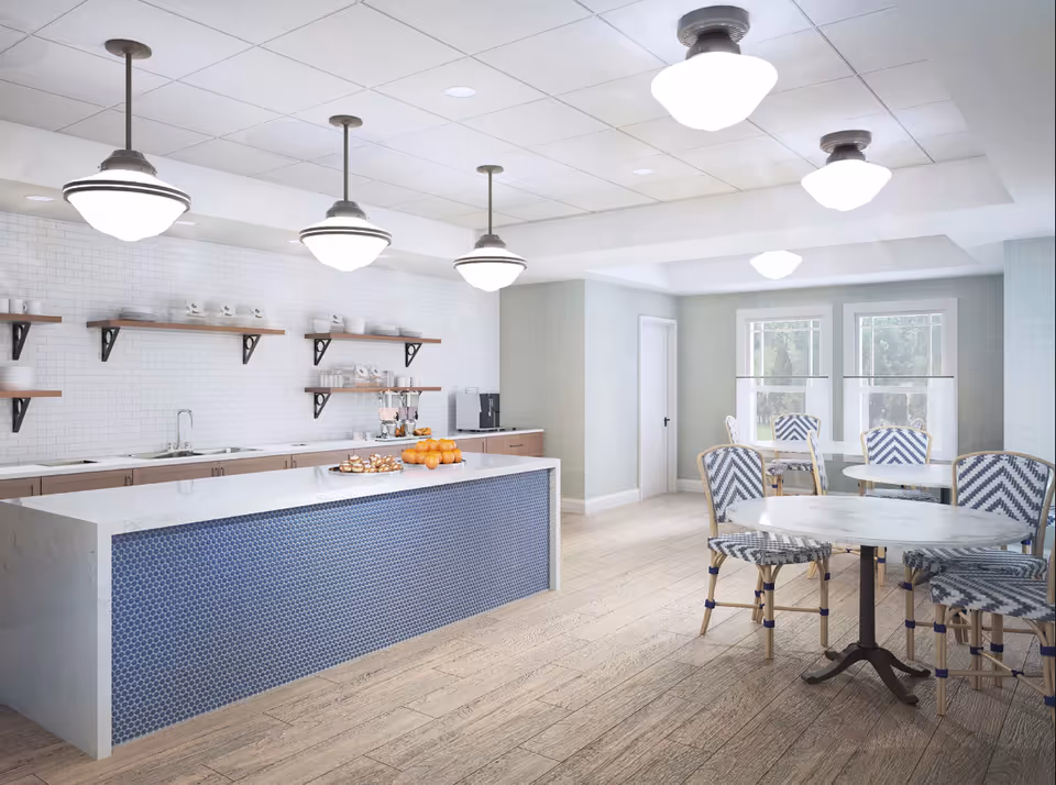 Bright communal kitchen and dining area with a long island, pendant lights, and round tables with patterned chairs.