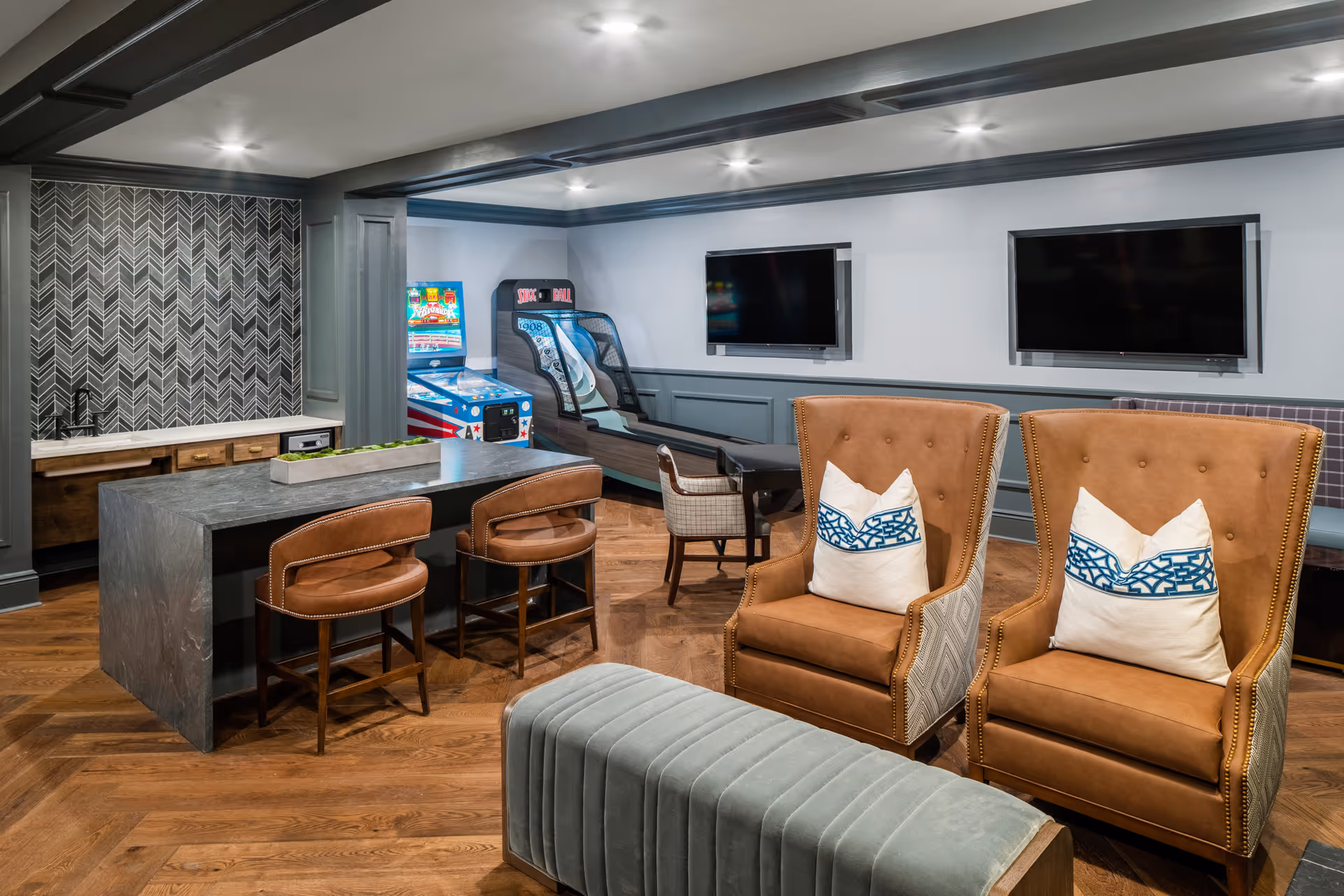 A cozy recreational room with two brown leather armchairs with white and blue patterned pillows, a gray upholstered bench, a dark stone table with two brown leather chairs, two wall-mounted flat-screen TVs, and arcade game machines in the background. The room features wood flooring and gray paneled walls with recessed lighting.