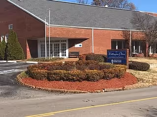Exterior view of a single-story brick building with a gray roof, surrounded by landscaped bushes and mulch. A blue sign in front reads 'Wellington Place Independent Living' with additional smaller text. The scene is set on a clear day with a paved road in the foreground.