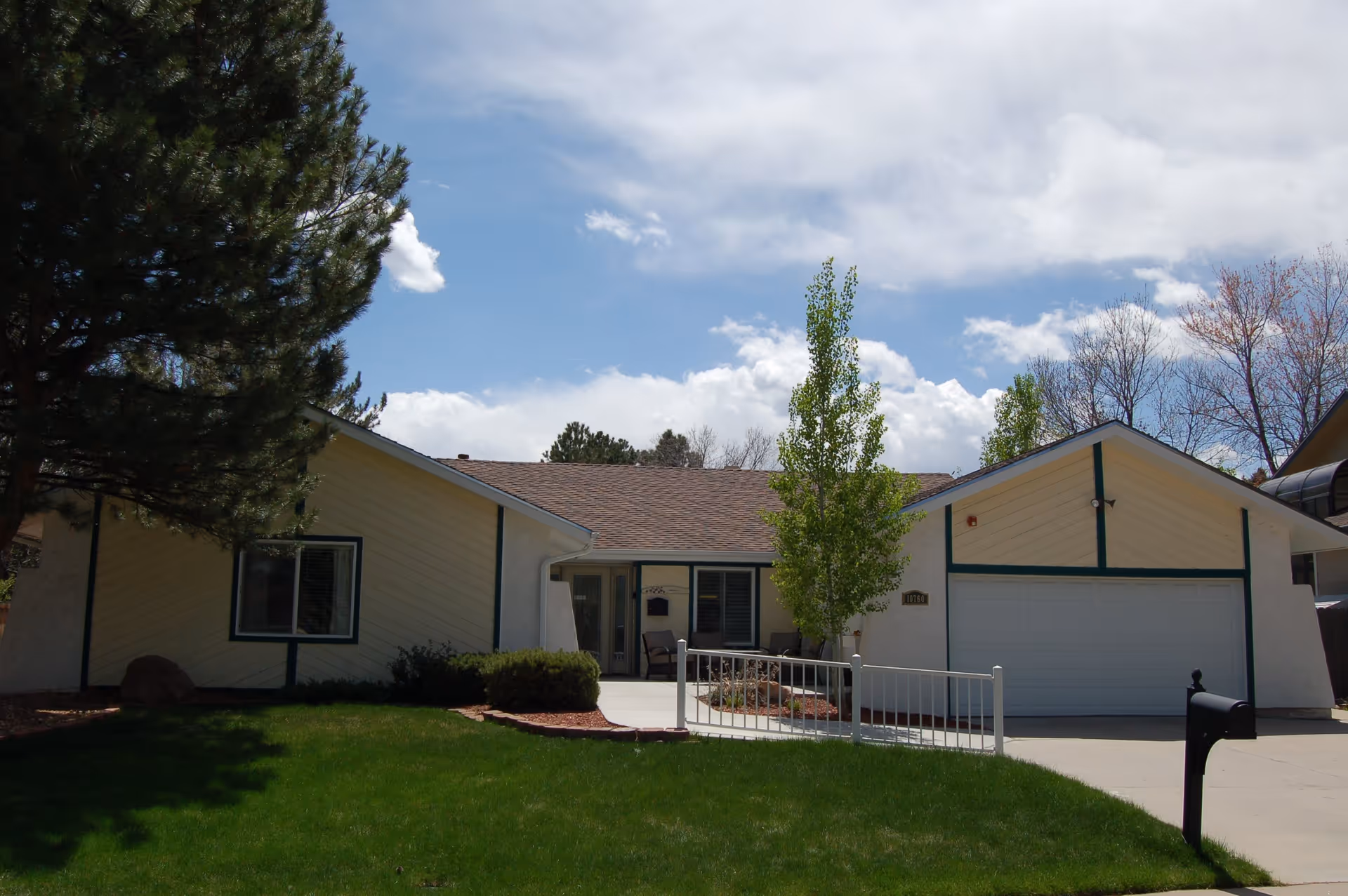 Single-story residential building with a light beige exterior and green trim, a two-car garage, a small front porch with chairs, a green lawn, a tree, and a clear blue sky with some clouds.