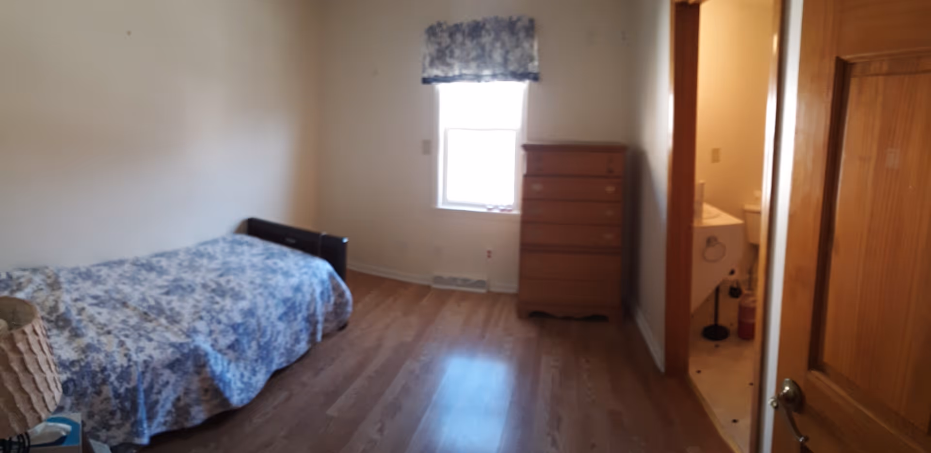 A small bedroom with a single bed covered in a blue floral bedspread on the left side, a wooden chest of drawers against the far wall under a window with a matching blue floral valance, and a doorway on the right leading to a bathroom with a sink and toilet visible.