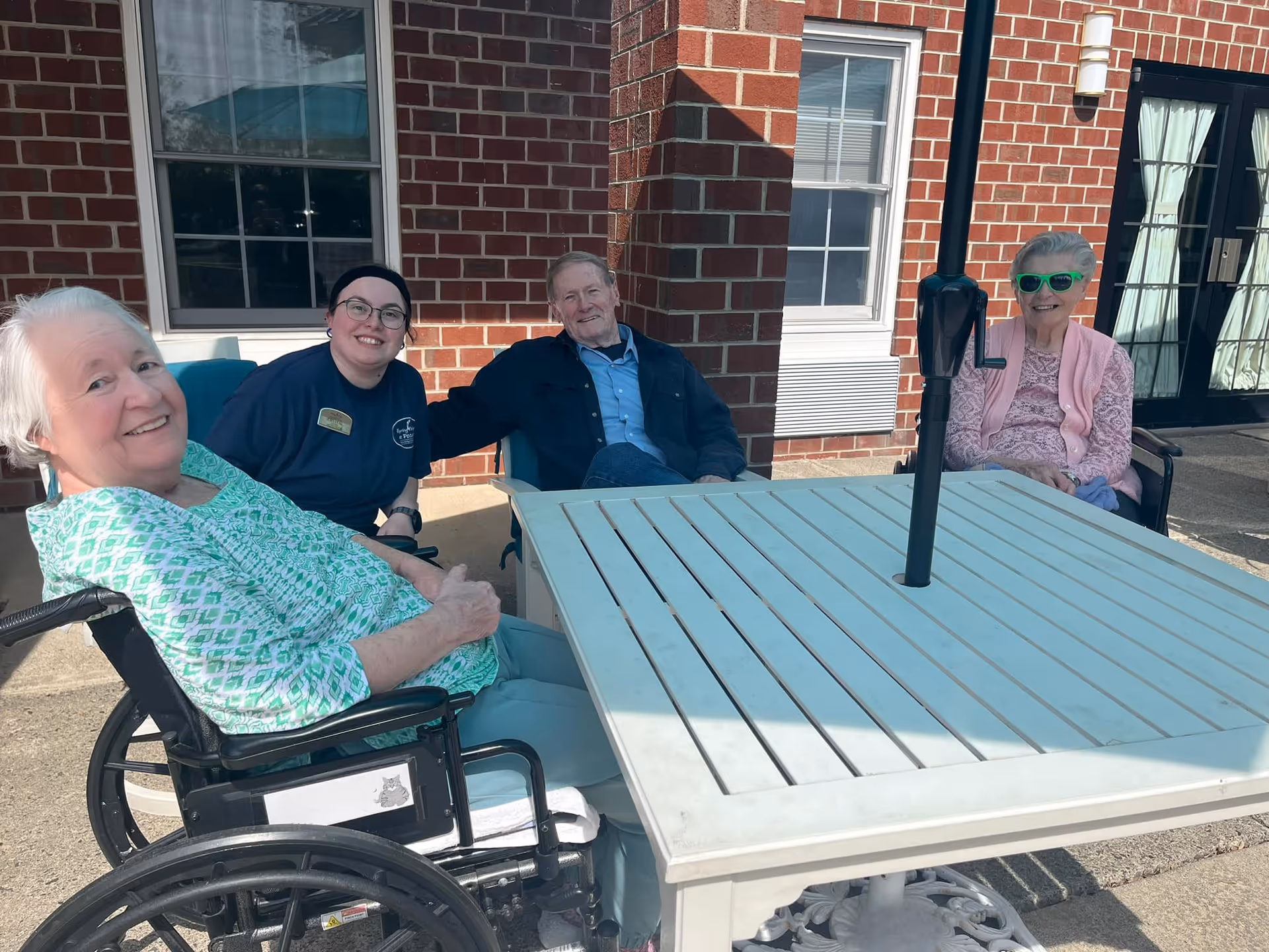 Four elderly individuals sitting around a white outdoor table with an umbrella pole in the center. They are outside a brick building with windows and a door with glass panels. One woman is in a wheelchair, another woman is wearing green sunglasses, and a younger person is sitting among them, smiling.