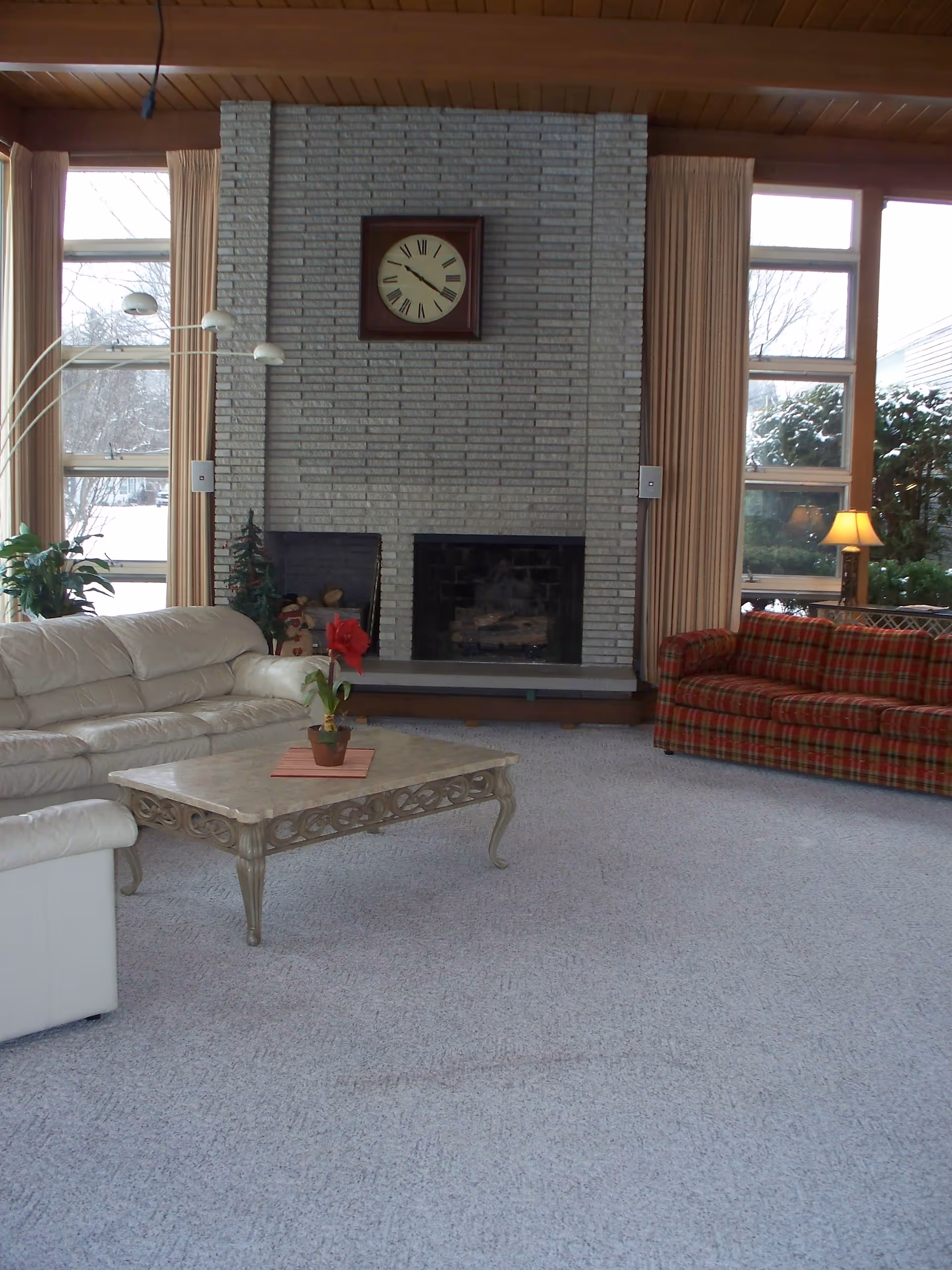 A spacious living room with a large brick fireplace in the center. Above the fireplace is a square clock with Roman numerals. The room has large windows with beige curtains on either side of the fireplace, letting in natural light. There is a white leather sofa on the left, a red plaid sofa on the right, and a decorative coffee table with a potted plant in the middle. A floor lamp with multiple lights is positioned near the white sofa, and a small lamp is visible on a side table near the red sofa. The carpet is light gray, and the ceiling has wooden paneling.