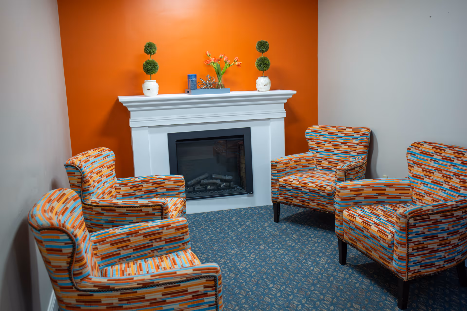 Cozy sitting area with four patterned armchairs arranged around a white fireplace against an orange accent wall.