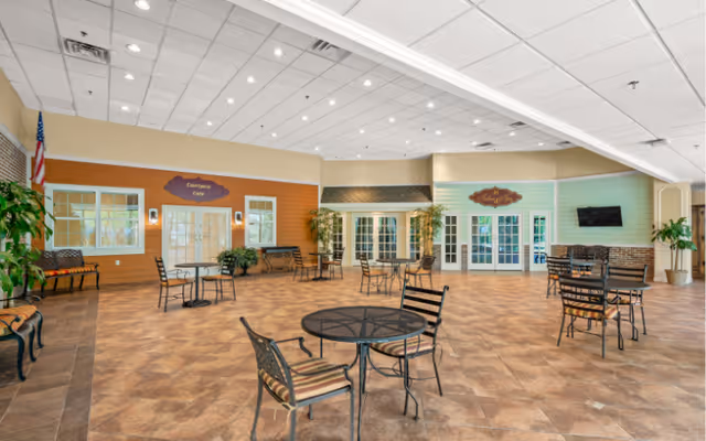Spacious indoor dining area with multiple round tables and chairs arranged on a tiled floor. The walls are painted in warm tones with signs indicating 'Everyday Cafe' and 'Market'. There are large windows and glass doors, potted plants, and an American flag in the corner. The ceiling has recessed lighting and a grid pattern.