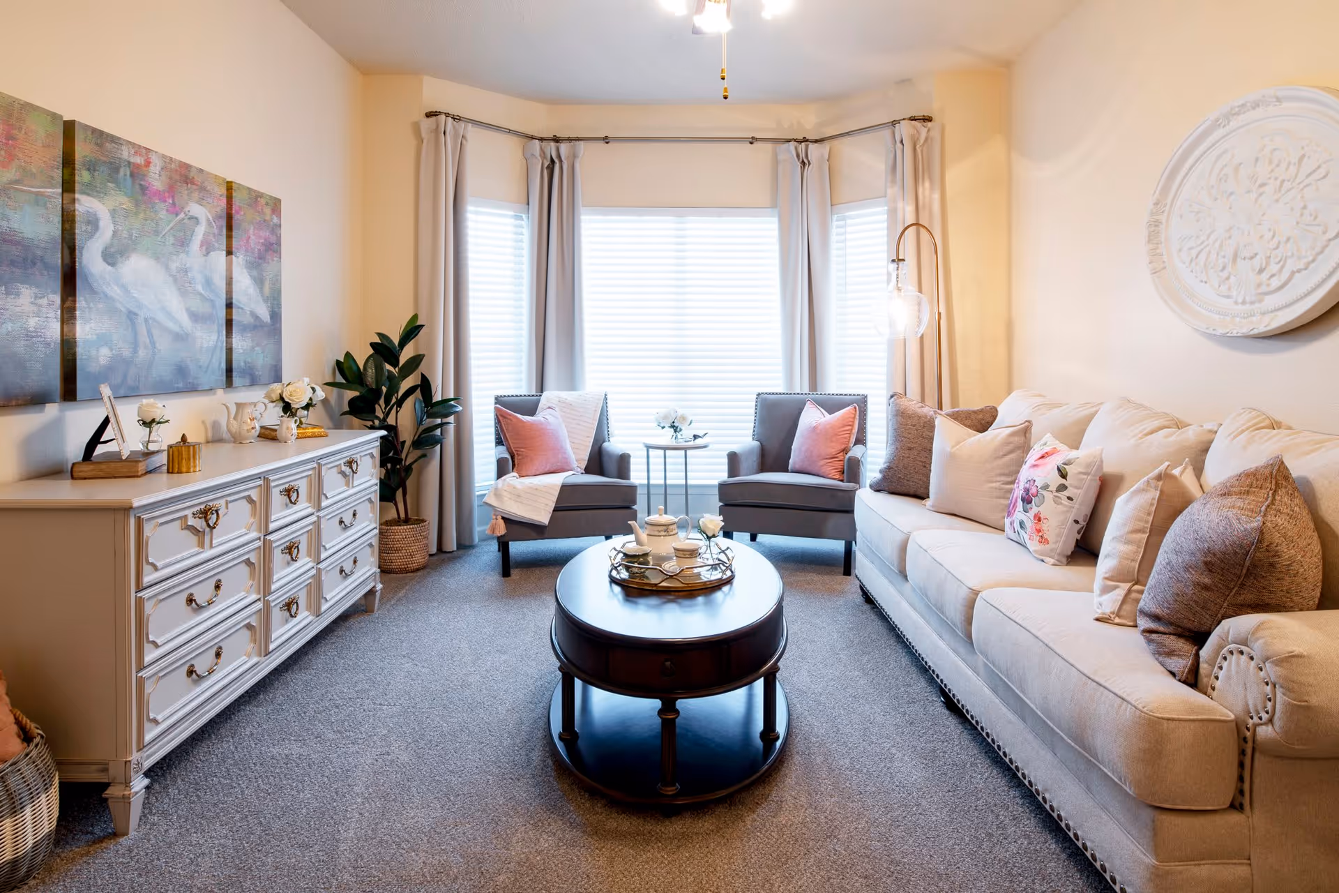 A cozy living room with a beige sofa adorned with multiple pillows, two gray armchairs with pink cushions, a round wooden coffee table with decorative items, a white dresser with ornate handles, a large plant in a woven basket, and a three-panel wall art featuring white birds. The room has large windows with white blinds and light-colored curtains, and a ceiling fan with a light fixture.