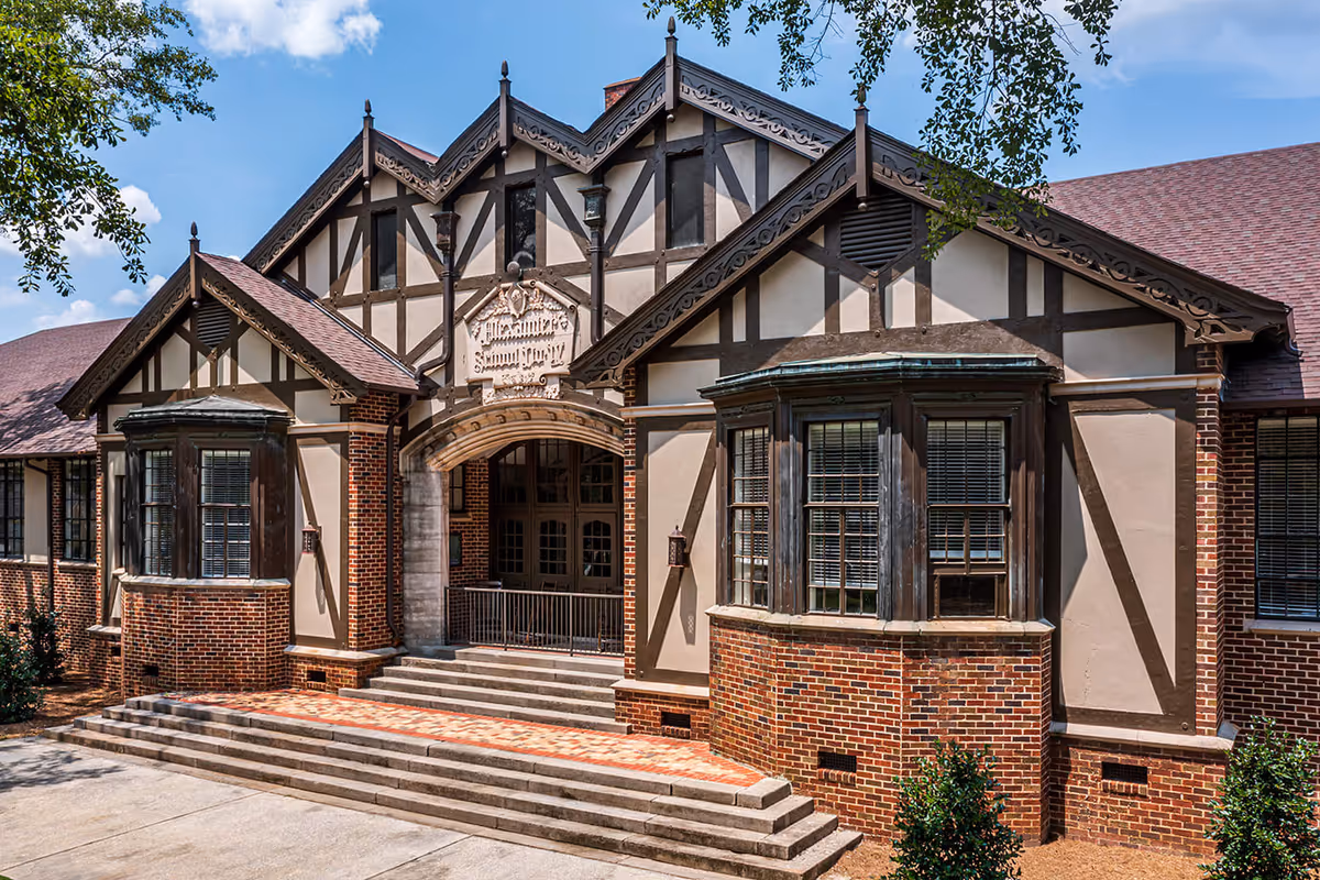 Front exterior view of a Tudor-style building with brick and timber framing, featuring a central arched entrance with steps leading up to double doors, flanked by bay windows and decorative wooden trim under a partly cloudy sky.