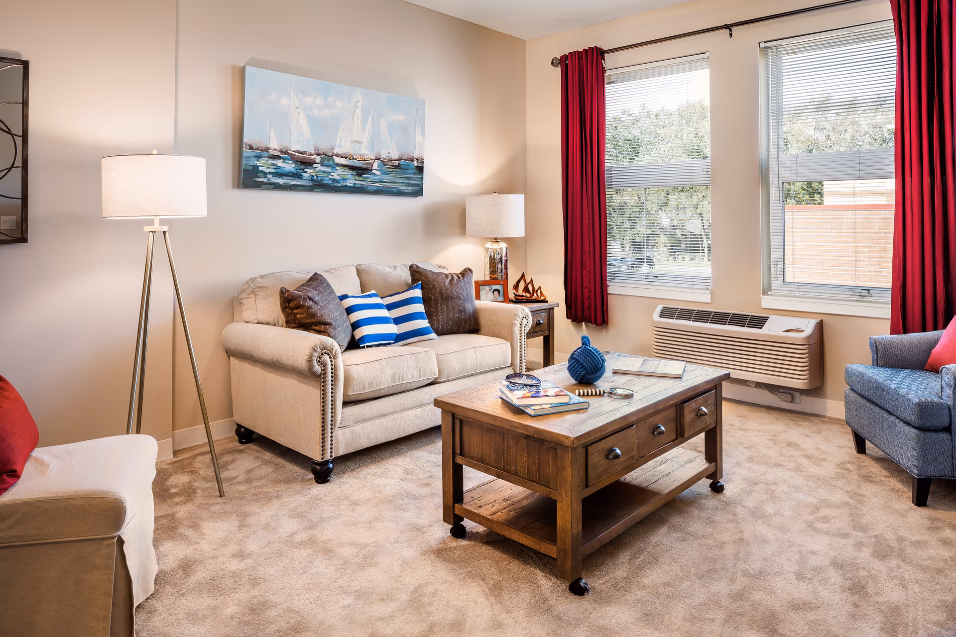 A cozy living room with a beige sofa adorned with blue and brown pillows, a wooden coffee table with books and decorative items, a floor lamp, a side table with a lamp and small decorations, two large windows with red curtains, and a blue armchair with a red pillow. The walls are light-colored and there is a painting of sailboats above the sofa.