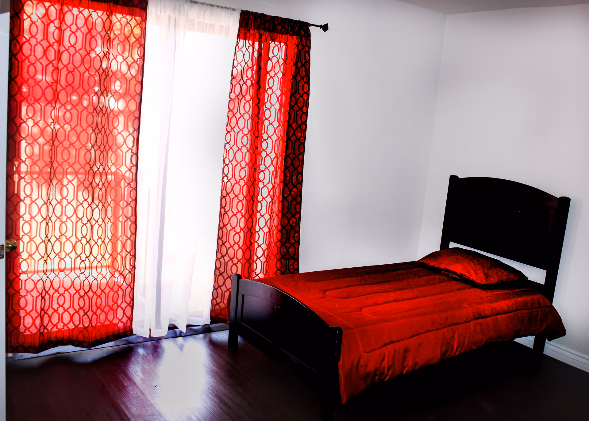 A simple bedroom with a single dark wooden bed frame, a red bedspread and matching pillow. The room has white walls and dark wooden flooring. There are large windows covered with red patterned curtains and sheer white curtains allowing light to filter in.