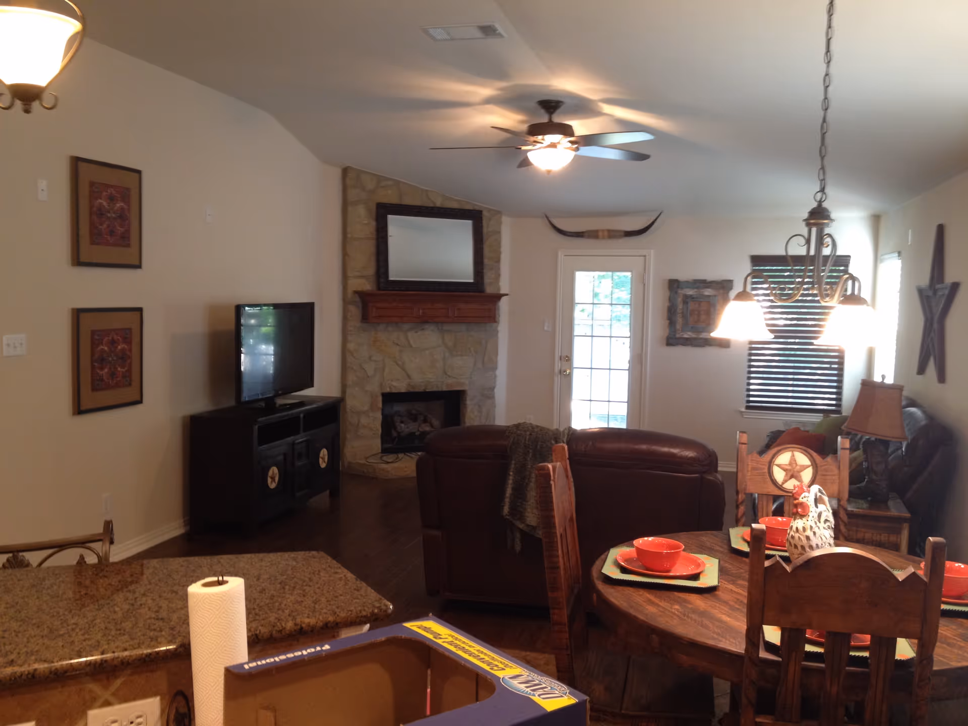 Interior view of a cozy living and dining area with a stone fireplace, a ceiling fan, a TV on a stand, a round wooden dining table set with red bowls and plates, and a leather couch. The room has warm lighting from ceiling fixtures and a window with blinds.