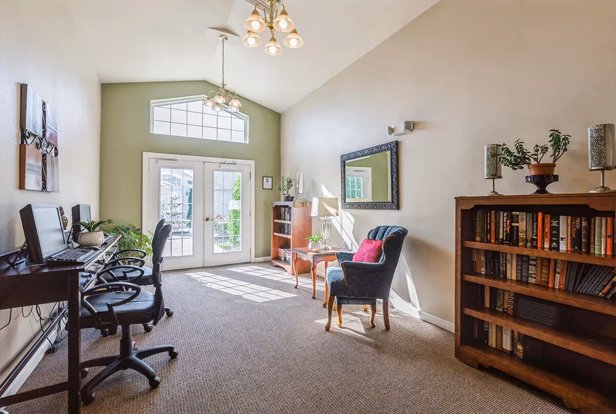 A bright room with a high ceiling featuring two chandeliers. On the left side, there are two computer desks with office chairs and plants. At the far end, there are double glass doors with a large window above, letting in natural light. On the right side, there is a blue upholstered armchair with a red cushion, a small wooden table with a lamp and plant, a mirror on the wall, and a wooden bookshelf filled with books and decorative items.