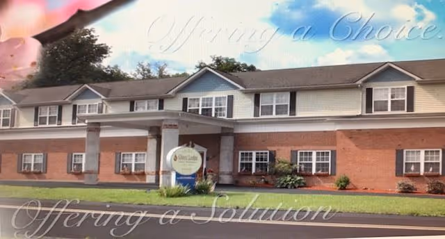 Exterior view of a two-story senior living facility building with a brick lower level and light-colored upper level. The building has multiple windows and a covered entrance supported by columns. There is a sign in front of the entrance that reads 'Gluco Lodge Personal Care Home'. The sky is partly cloudy.