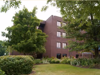 Brick multi-story building with windows set behind trees and landscaped shrubs and lawn.