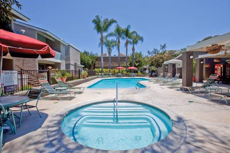 Outdoor pool area at Heritage Village Anaheim For Seniors featuring a small circular hot tub in the foreground, a larger rectangular swimming pool in the background, surrounded by lounge chairs, tables with umbrellas, palm trees, and residential buildings under a clear blue sky.