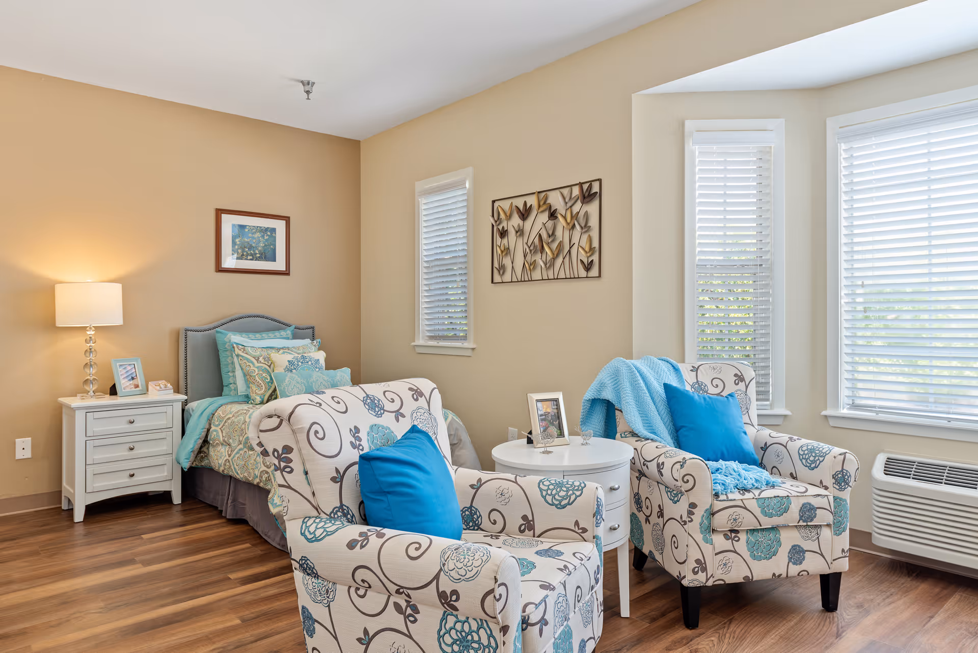 A cozy bedroom in an assisted living facility featuring a single bed with teal and patterned bedding, a white nightstand with a lamp and framed photos, two patterned armchairs with blue cushions and a blue throw blanket, a small round white table between the chairs, and windows with white blinds letting in natural light.