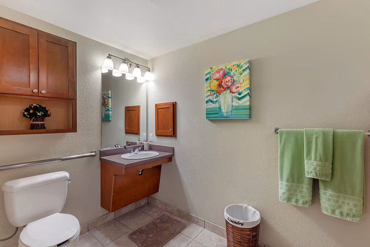 A clean bathroom with a white toilet, a wall-mounted wooden cabinet, a sink with a wooden vanity, a large mirror above the sink, and a light fixture with four bulbs. There is a colorful floral painting on the wall and two green towels hanging on a towel rack. A wicker laundry basket is placed on the tiled floor.